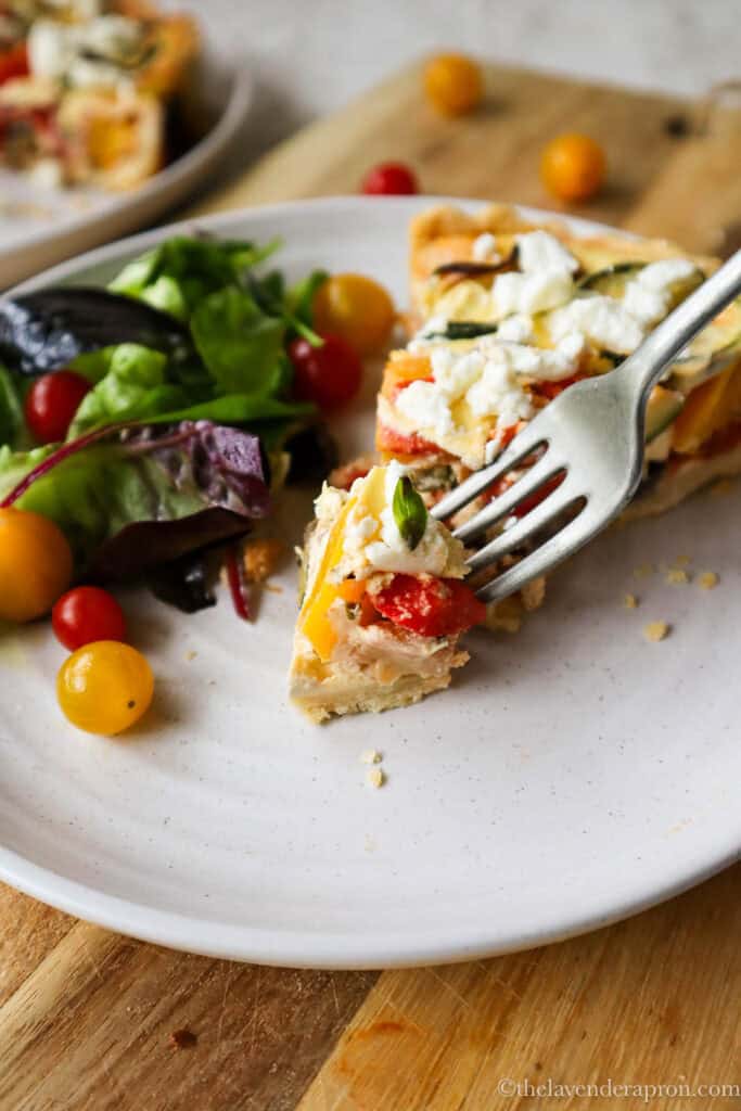 A forkful of ratatouille quiche showing yellow and red bell pepper, tomato, zucchini and eggplant and a side garden salad.