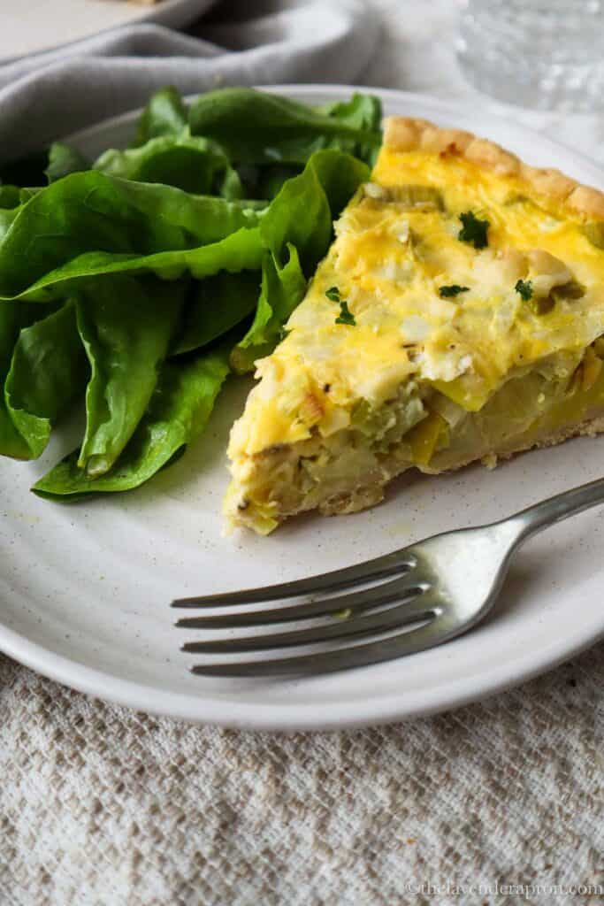 Slice of leek and gruyère quiche on a plate with a green salad and fork.