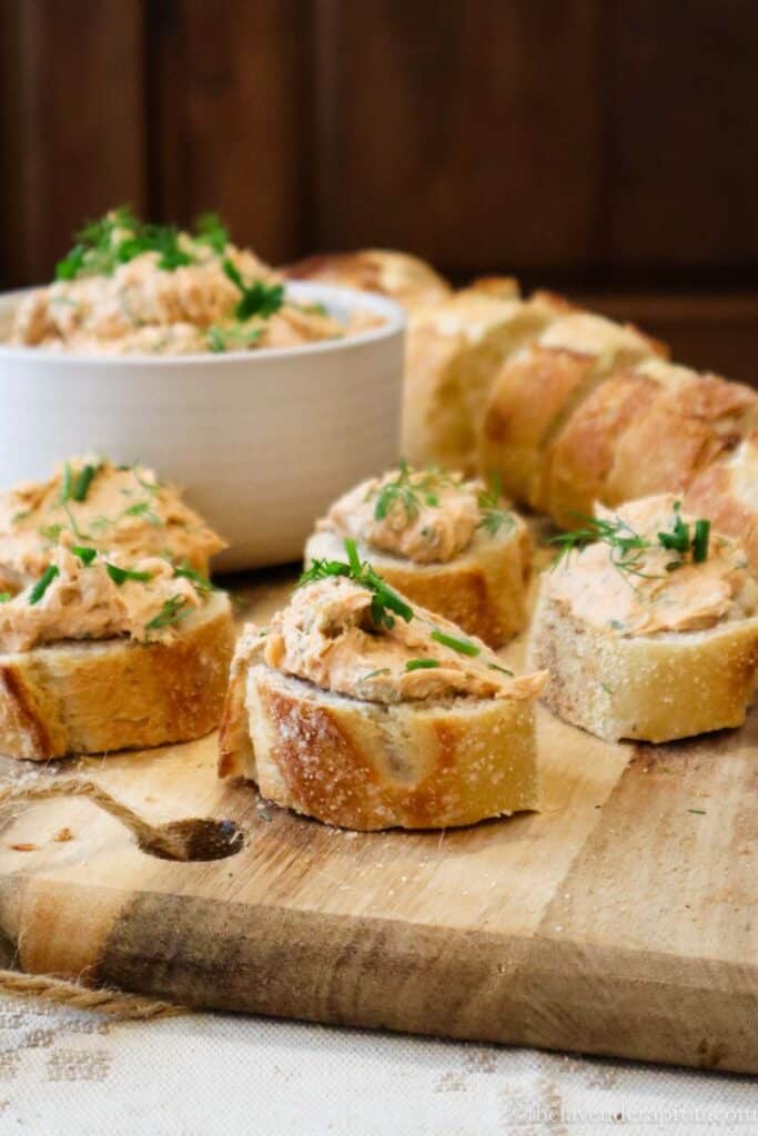 Close up of salmon cream cheese spread on slice of toast. There are 5 toasts on a wooden cutting board and a small bowl of the mixture in the background, along with slices of baguette toasts.