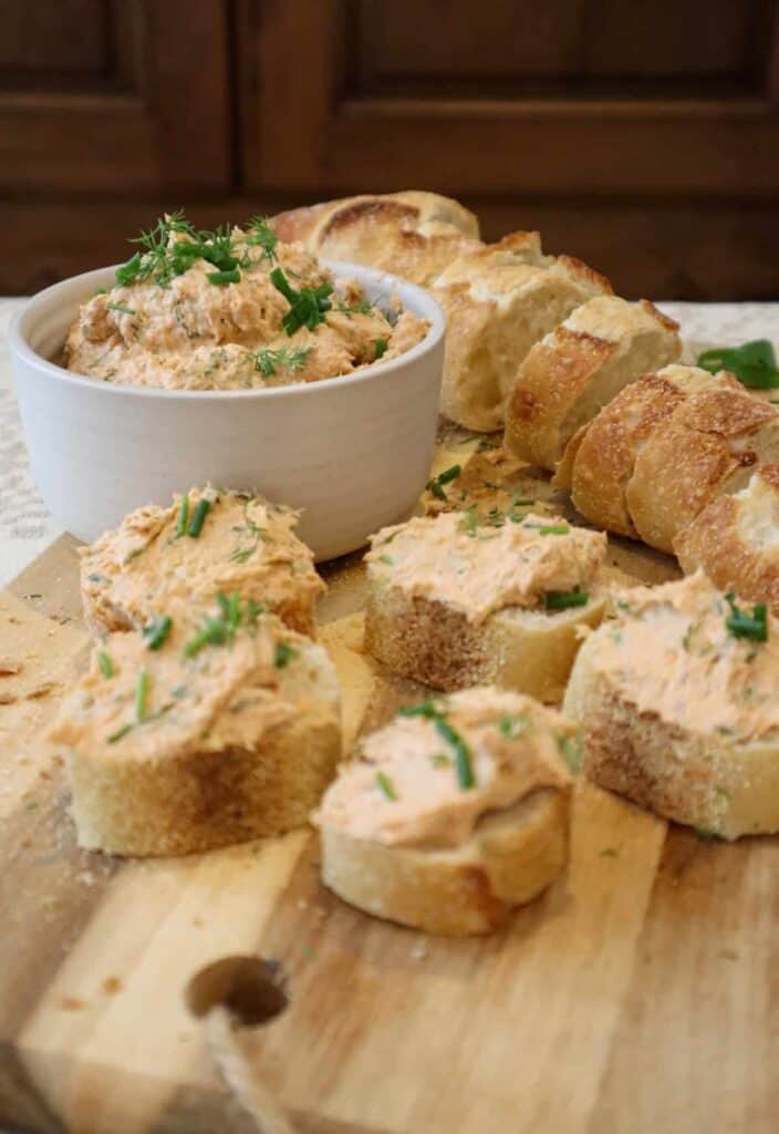 Canned salmon and cream cheese spread in small bowl placed on wooden cutting board, along with slices of sourdough baguette and five salmon cream cheese toasts topped with chopped fresh dill, chives and parsley