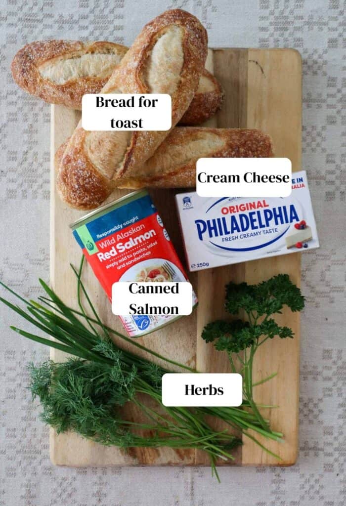 Sourdough bread, canned red salmon, fresh dill, chives, spring onion and parsley and cream cheese all placed on a wooden chopping board, which is placed on a linen napkin.