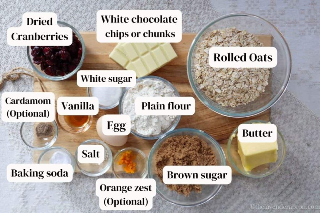 Traditional oats, dried cranberries, white chocolate bar, flour, brown sugar, white sugar, vanilla, cardamom, salt, butter on a wooden board.